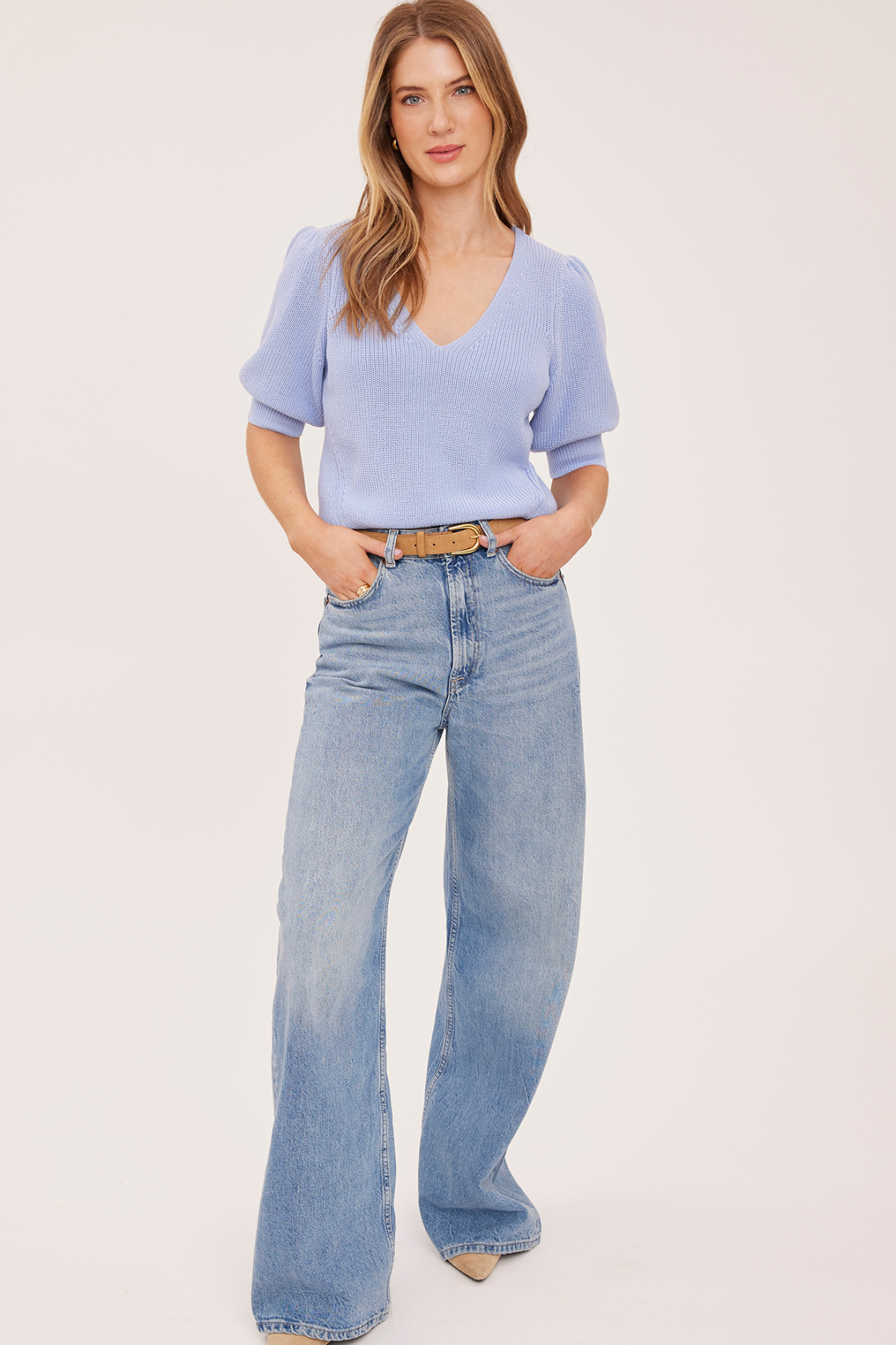 A woman modeling a light blue ribbed knit short-sleeve V-neck top paired with light-wash wide-leg jeans and a tan belt, standing against a plain light beige studio background.