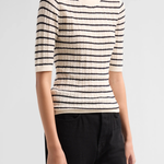 A woman wearing a cream-colored short-sleeve knit sweater with thin black horizontal stripes and black pants, photographed against a light gray studio background.