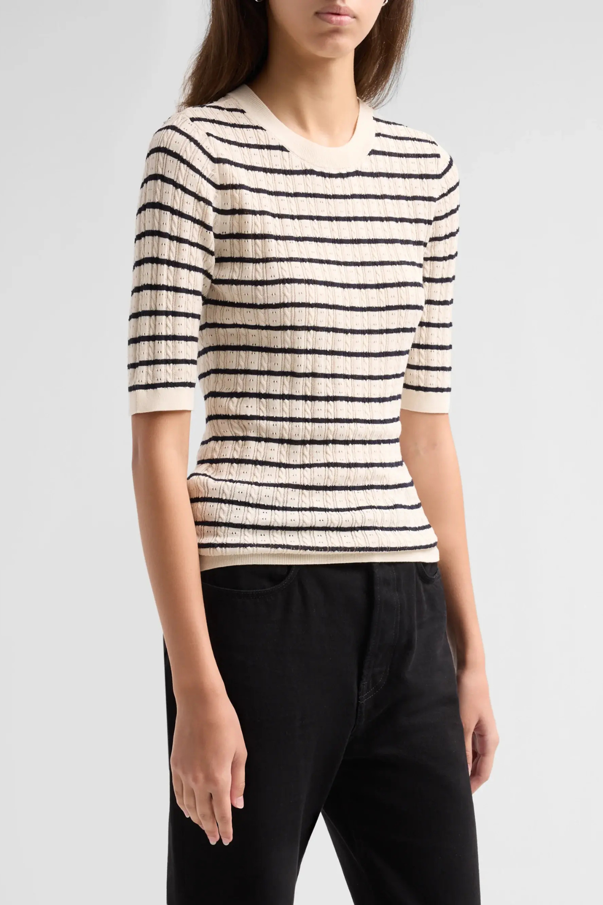 A woman wearing a cream-colored short-sleeve knit sweater with thin black horizontal stripes and black pants, photographed against a light gray studio background.