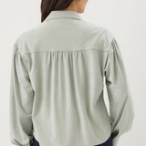A woman viewed from the back wearing a light sage green long-sleeve blouse with gathered detailing at the shoulders and dark blue jeans, set against a plain white studio background.