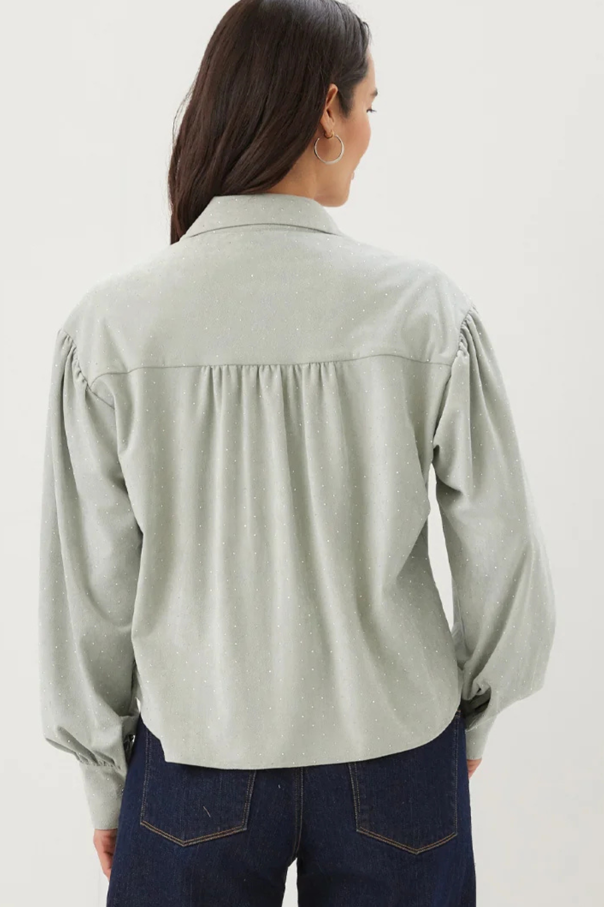 A woman viewed from the back wearing a light sage green long-sleeve blouse with gathered detailing at the shoulders and dark blue jeans, set against a plain white studio background.