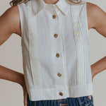 Person wearing a white sleeveless button-up top with pleated details and blue jeans.