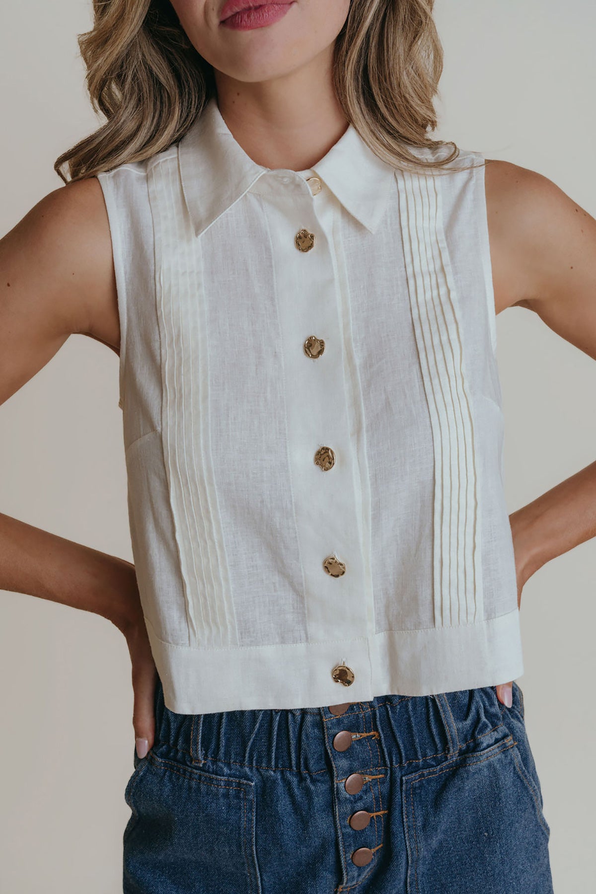 Person wearing a white sleeveless button-up top with pleated details and blue jeans.