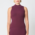 A woman modeling a sleeveless ribbed knit mock neck dress in a deep plum purple color, paired with gold hoop earrings, against a light gray studio background.