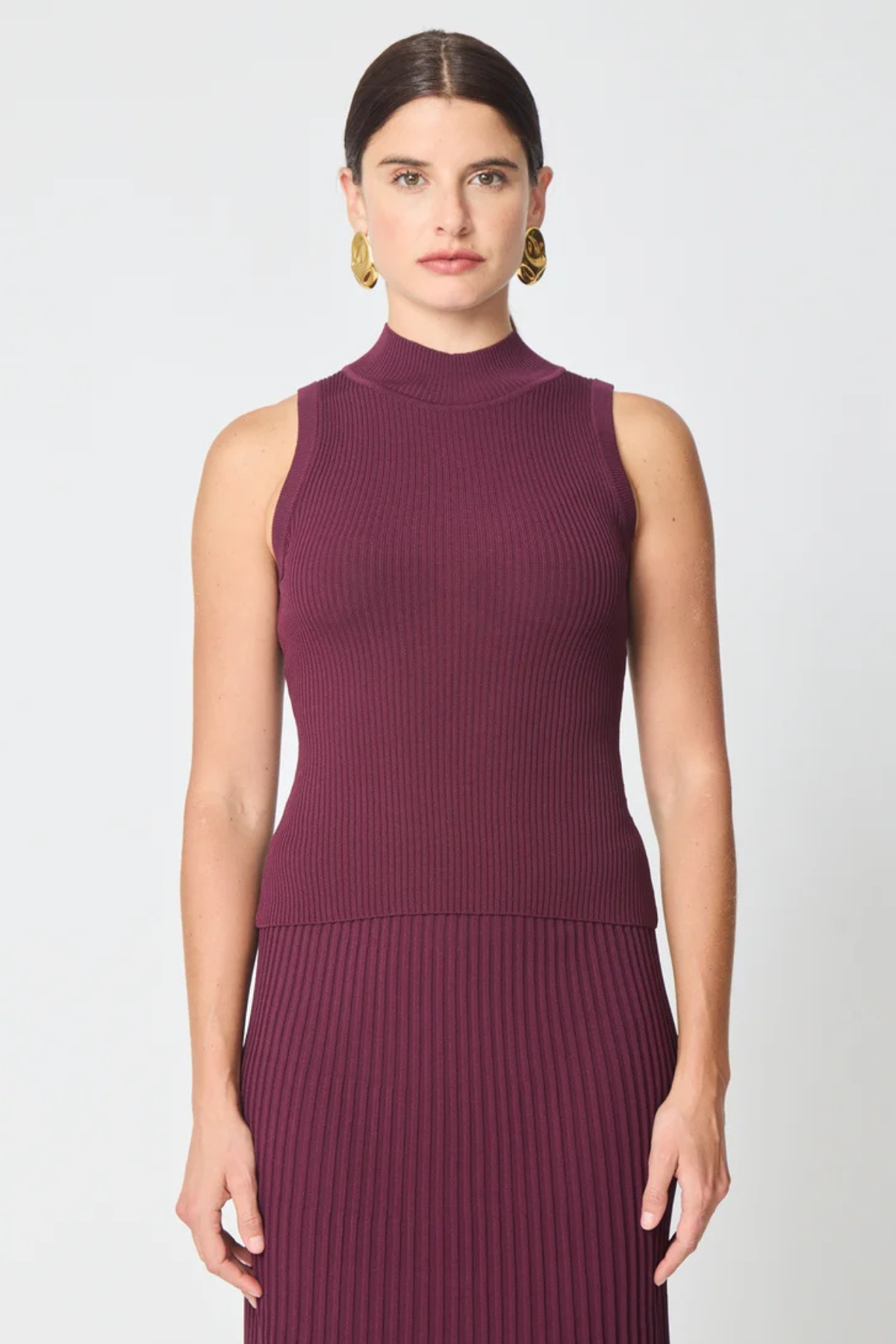 A woman modeling a sleeveless ribbed knit mock neck dress in a deep plum purple color, paired with gold hoop earrings, against a light gray studio background.