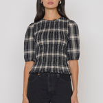 A woman modeling a black and beige plaid smocked top with puff sleeves, paired with black jeans, against a plain light gray background.
