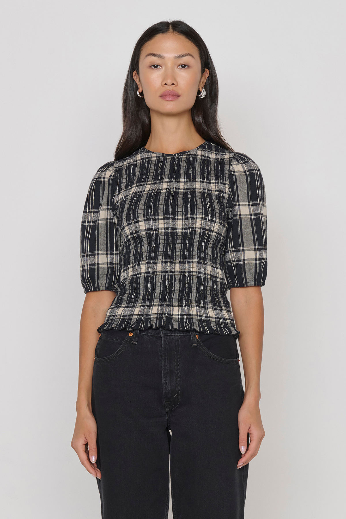 A woman modeling a black and beige plaid smocked top with puff sleeves, paired with black jeans, against a plain light gray background.