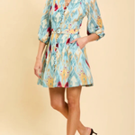 A woman models a light blue and yellow patterned mini shirt dress with a matching belt, paired with black heels, against a solid beige background.
