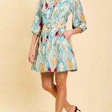 A woman models a light blue and yellow patterned mini shirt dress with a matching belt, paired with black heels, against a solid beige background.