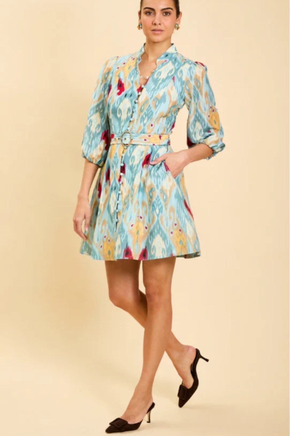 A woman models a light blue and yellow patterned mini shirt dress with a matching belt, paired with black heels, against a solid beige background.