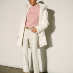 A model stands against a white wall wearing a white oversized coat over a light pink mock-neck top, white trousers, and beige pointed-toe ankle boots.