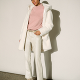 A model stands against a white wall wearing a white oversized coat over a light pink mock-neck top, white trousers, and beige pointed-toe ankle boots.