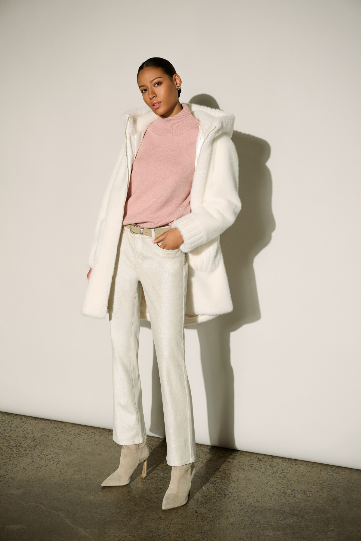 A model stands against a white wall wearing a white oversized coat over a light pink mock-neck top, white trousers, and beige pointed-toe ankle boots.