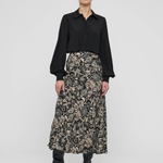 A model stands against a light gray studio background wearing a black long-sleeve button-up shirt and a high-waisted midi skirt with a beige and black floral pattern, paired with black pointed-toe heeled boots.