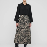 A model stands against a light gray studio background wearing a black long-sleeve button-up shirt and a high-waisted midi skirt with a beige and black floral pattern, paired with black pointed-toe heeled boots.
