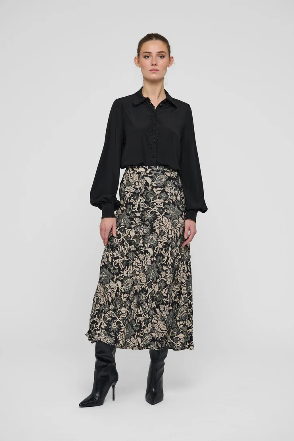 A model stands against a light gray studio background wearing a black long-sleeve button-up shirt and a high-waisted midi skirt with a beige and black floral pattern, paired with black pointed-toe heeled boots.