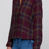 A woman wearing a long-sleeve button-up plaid shirt in burgundy with blue and yellow stripes, paired with blue jeans, against a plain light gray background.