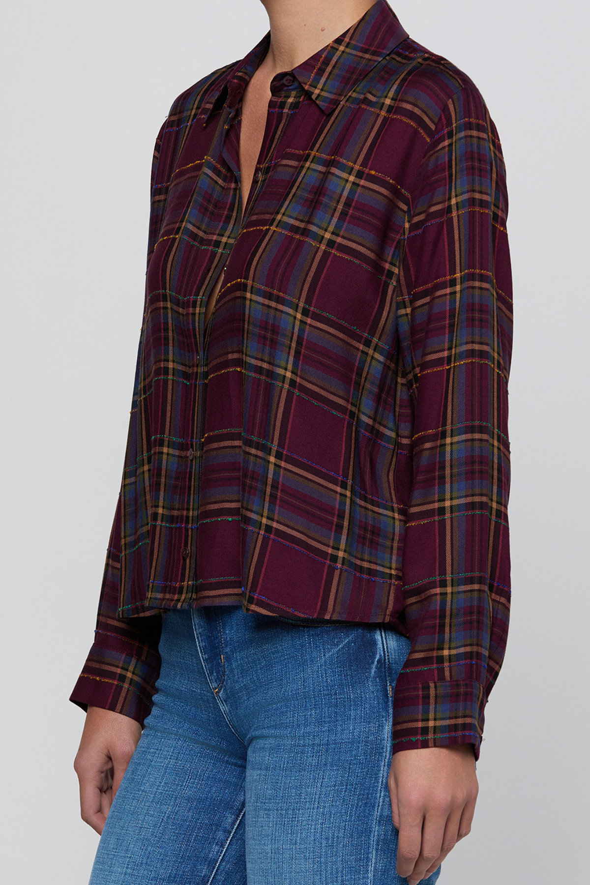 A woman wearing a long-sleeve button-up plaid shirt in burgundy with blue and yellow stripes, paired with blue jeans, against a plain light gray background.
