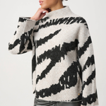 A woman wearing a fuzzy knit sweater with a black and off-white zebra-like pattern, paired with dark metallic pants, against a plain light gray studio background.