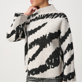 A woman wearing a fuzzy knit sweater with a black and off-white zebra-like pattern, paired with dark metallic pants, against a plain light gray studio background.