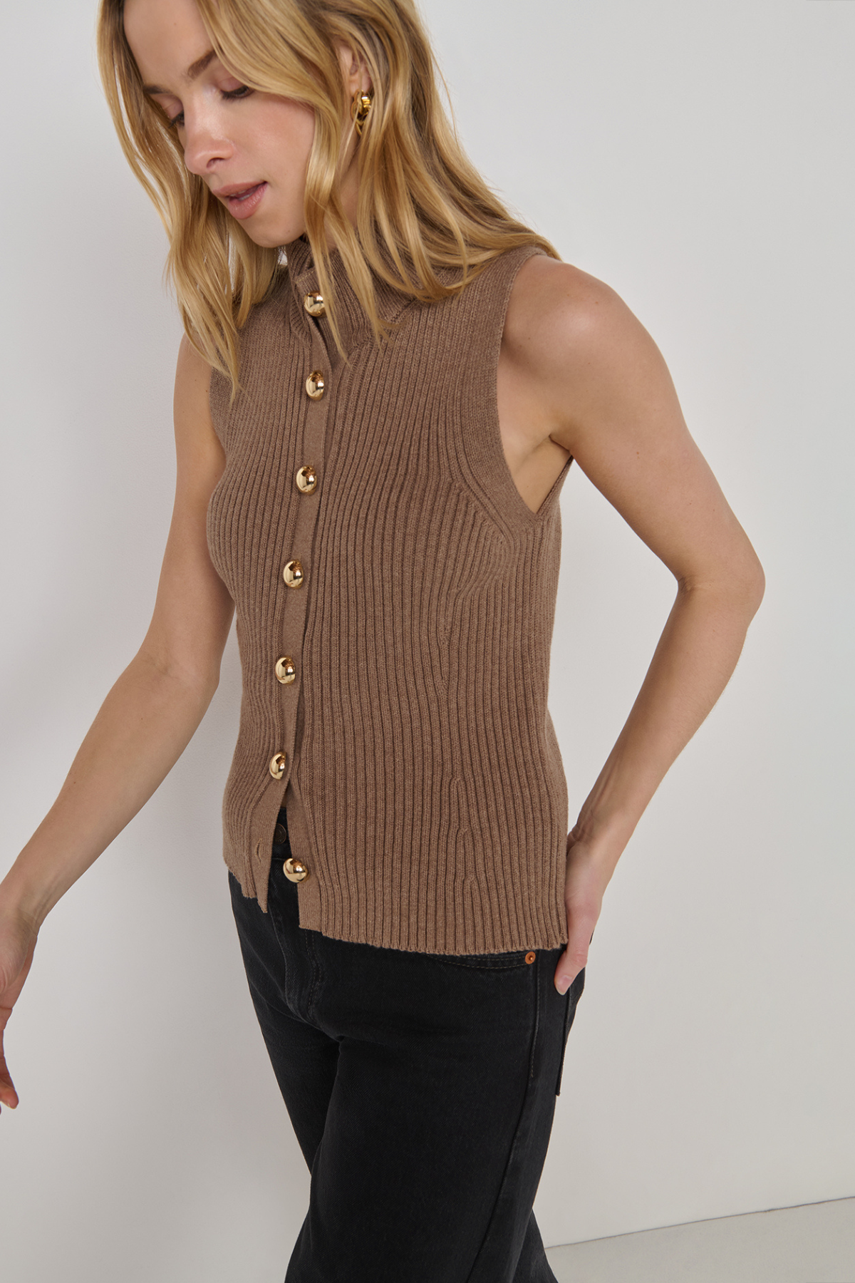 A woman wearing a sleeveless ribbed knit vest in taupe brown with gold-tone buttons, paired with black pants, photographed against a plain light gray studio background.