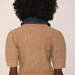 Back view of a person with a large black afro wearing a beige short-sleeve cable-knit sweater over a dark blue denim top, paired with brown velvet pants, against a plain off-white background.