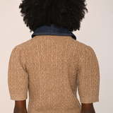 Back view of a person with a large black afro wearing a beige short-sleeve cable-knit sweater over a dark blue denim top, paired with brown velvet pants, against a plain off-white background.