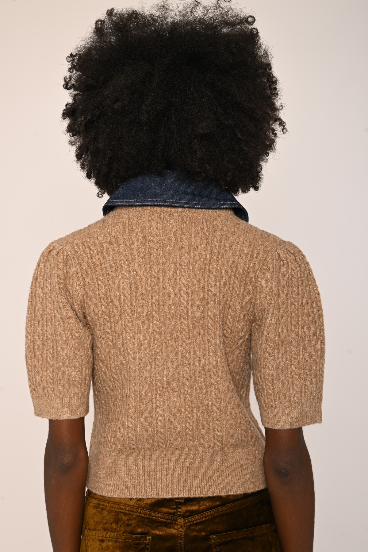 Back view of a person with a large black afro wearing a beige short-sleeve cable-knit sweater over a dark blue denim top, paired with brown velvet pants, against a plain off-white background.