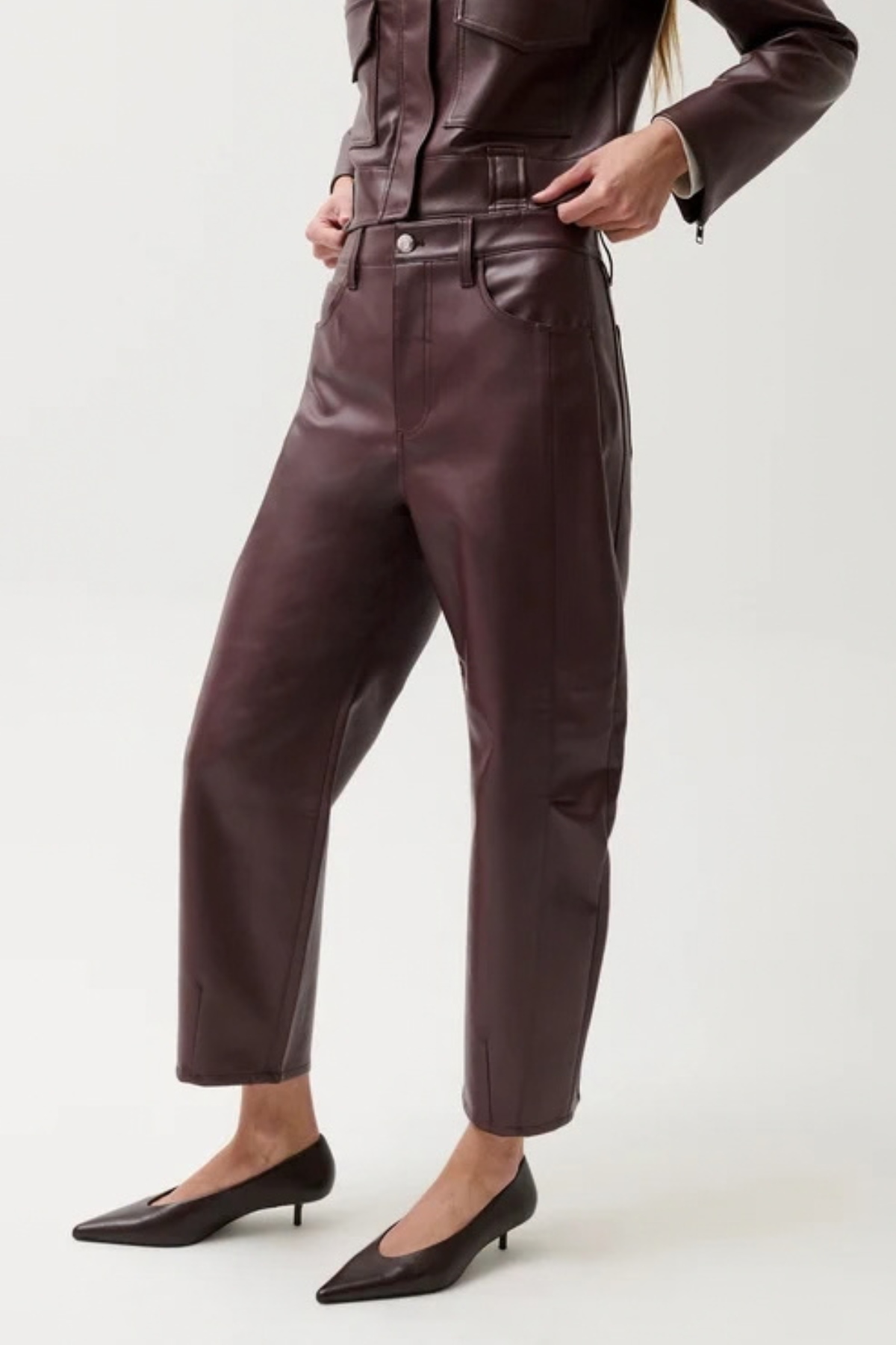 A model wearing dark brown faux leather cropped pants with a belt, paired with black pointed-toe kitten heels, shown from the waist down on a light gray studio background.