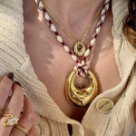 A close-up lifestyle shot of a model wearing a layered necklace set. The set features a large gold crescent moon pendant on a braided cream and burgundy cord, layered over a gold chain with a small gold medallion, and a gold ring on the model's finger.