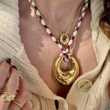 A close-up lifestyle shot of a model wearing a layered necklace set. The set features a large gold crescent moon pendant on a braided cream and burgundy cord, layered over a gold chain with a small gold medallion, and a gold ring on the model's finger.