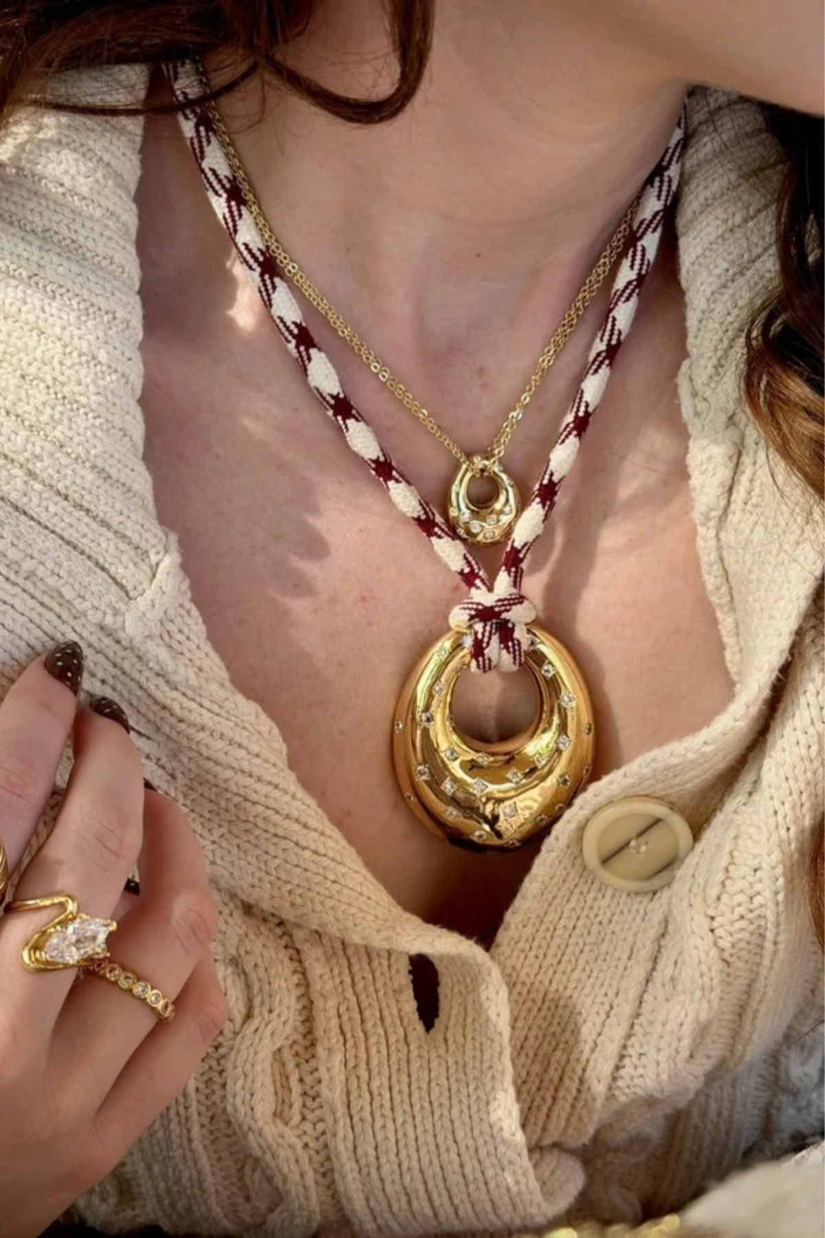 A close-up lifestyle shot of a model wearing a layered necklace set. The set features a large gold crescent moon pendant on a braided cream and burgundy cord, layered over a gold chain with a small gold medallion, and a gold ring on the model's finger.