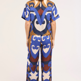 A woman viewed from the back wearing a matching short-sleeve top and wide-leg pants set with a bold abstract pattern in blue, brown, cream, and white on a light studio background.