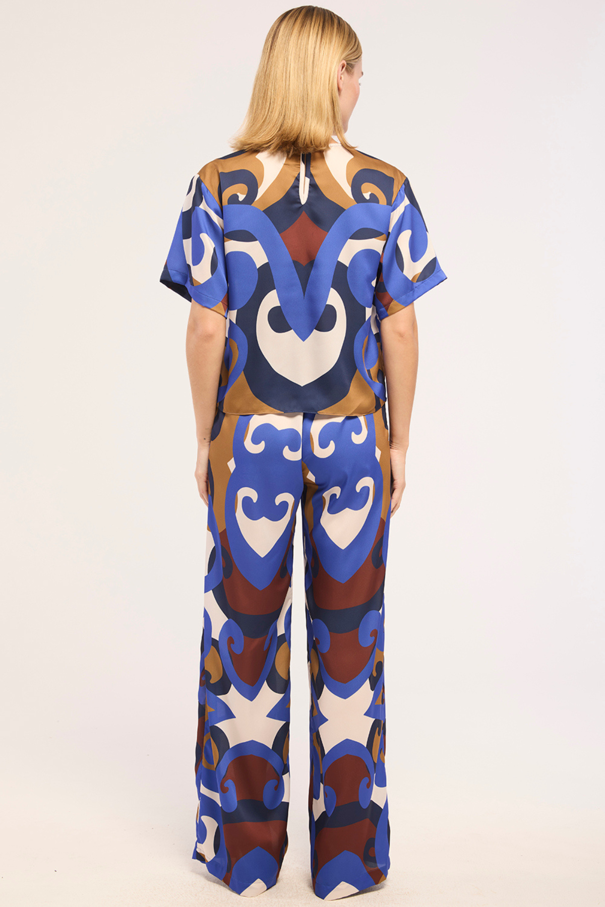 A woman viewed from the back wearing a matching short-sleeve top and wide-leg pants set with a bold abstract pattern in blue, brown, cream, and white on a light studio background.