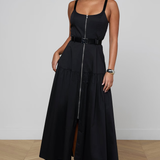 A woman modeling a black sleeveless maxi dress with a front zipper, a matching black belt at the waist, and a tiered skirt, standing against a light gray wall and wood floor.