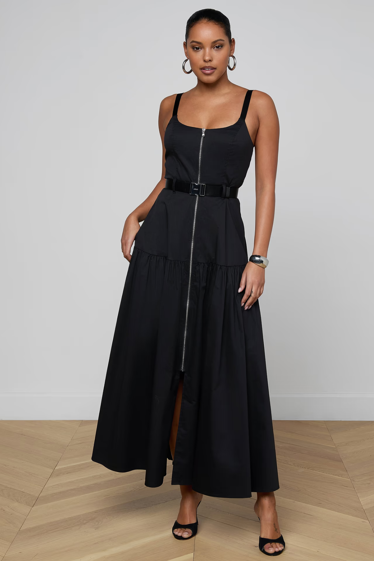 A woman modeling a black sleeveless maxi dress with a front zipper, a matching black belt at the waist, and a tiered skirt, standing against a light gray wall and wood floor.