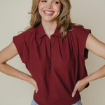 A smiling woman with blonde hair wearing a sleeveless, pleated burgundy polo shirt and light blue jeans, posing with her hands on her hips against a solid beige background.