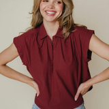 A smiling woman with blonde hair wearing a sleeveless, pleated burgundy polo shirt and light blue jeans, posing with her hands on her hips against a solid beige background.