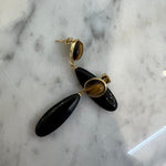 A pair of black teardrop dangle earrings with gold-toned posts and tiger's eye inlays, displayed on a white and gray marble surface.