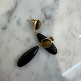 A pair of black teardrop dangle earrings with gold-toned posts and tiger's eye inlays, displayed on a white and gray marble surface.
