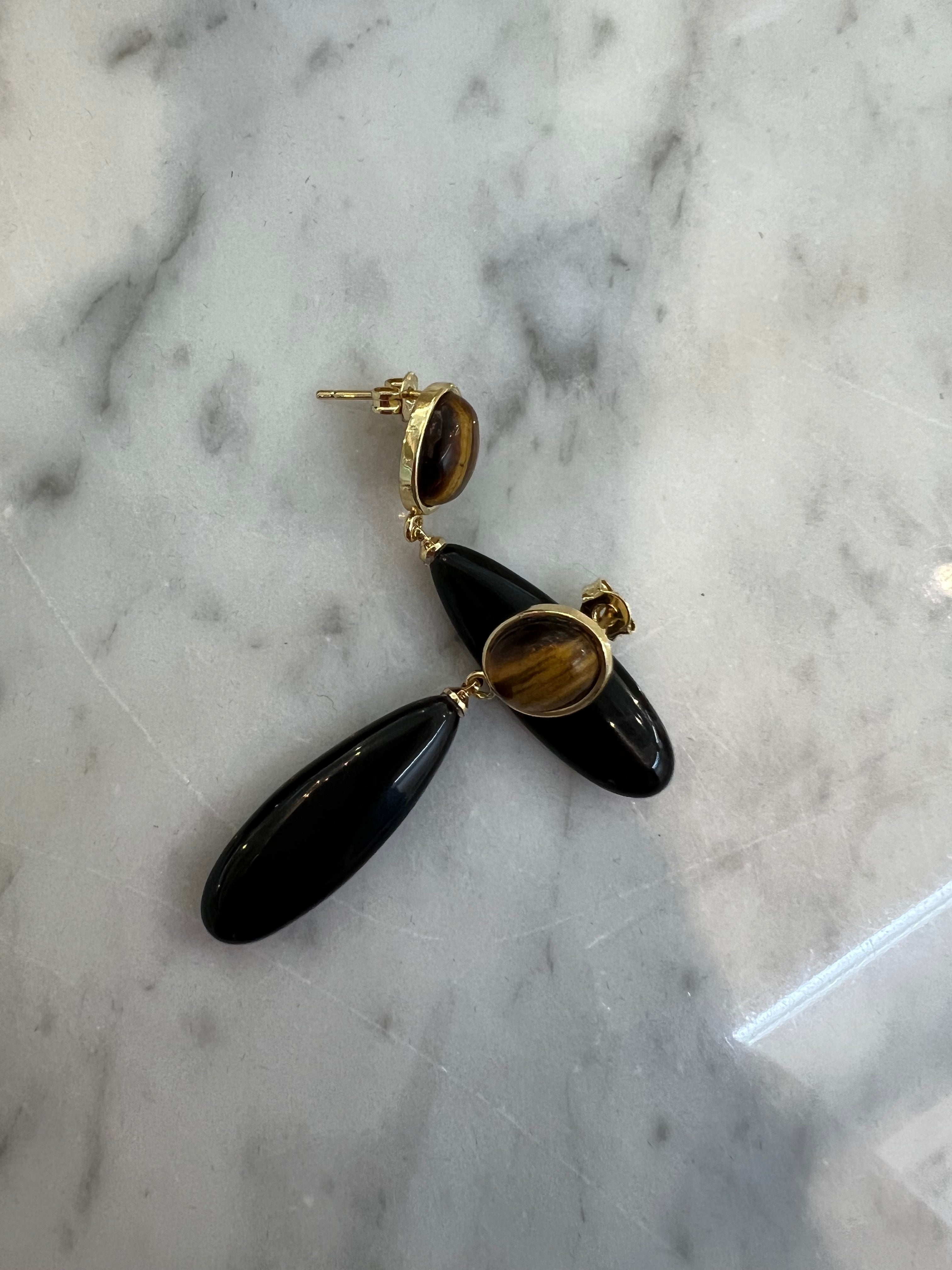 A pair of black teardrop dangle earrings with gold-toned posts and tiger's eye inlays, displayed on a white and gray marble surface.