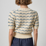 A woman shown from the back wearing a cream knit short-sleeve top with a blue wavy stripe pattern and a matching ribbed hem, paired with a dark blue skirt, against a light gray studio background.