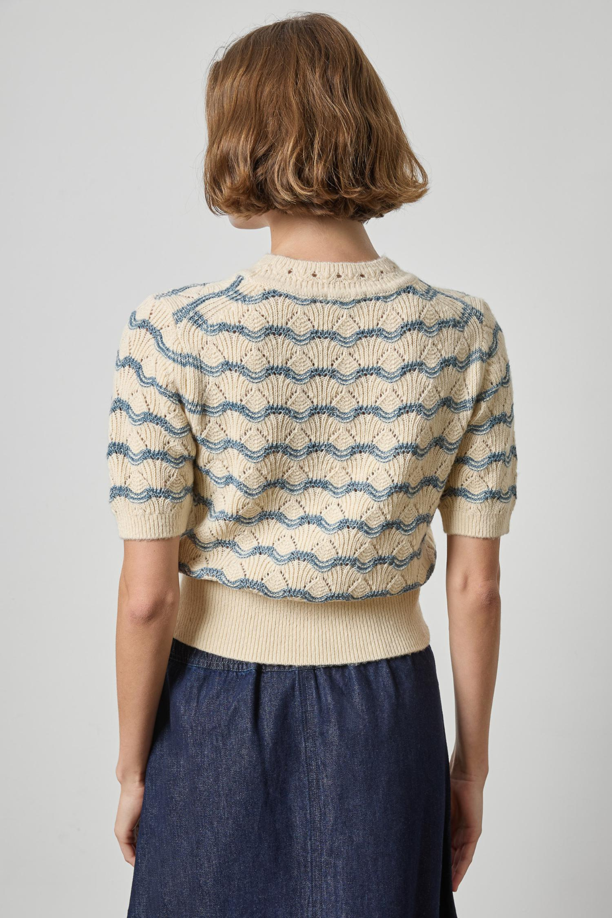 A woman shown from the back wearing a cream knit short-sleeve top with a blue wavy stripe pattern and a matching ribbed hem, paired with a dark blue skirt, against a light gray studio background.
