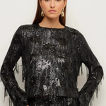A woman modeling a black long-sleeve top covered in shimmering sequins and fringe, paired with a matching fringed skirt, against a plain light background.