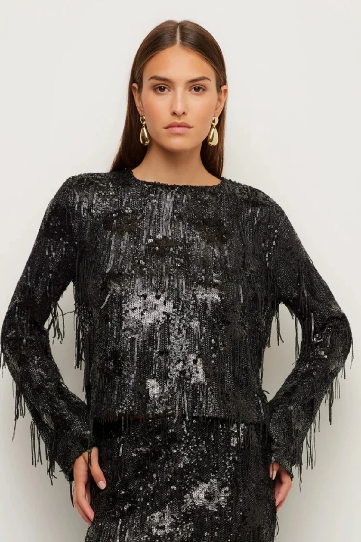 A woman modeling a black long-sleeve top covered in shimmering sequins and fringe, paired with a matching fringed skirt, against a plain light background.