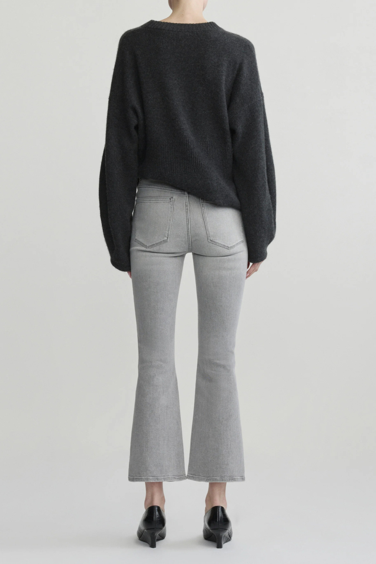 A model shown from the back wearing a dark charcoal gray knit sweater, light gray flared jeans, and black pointed-toe heels against a light gray studio background.