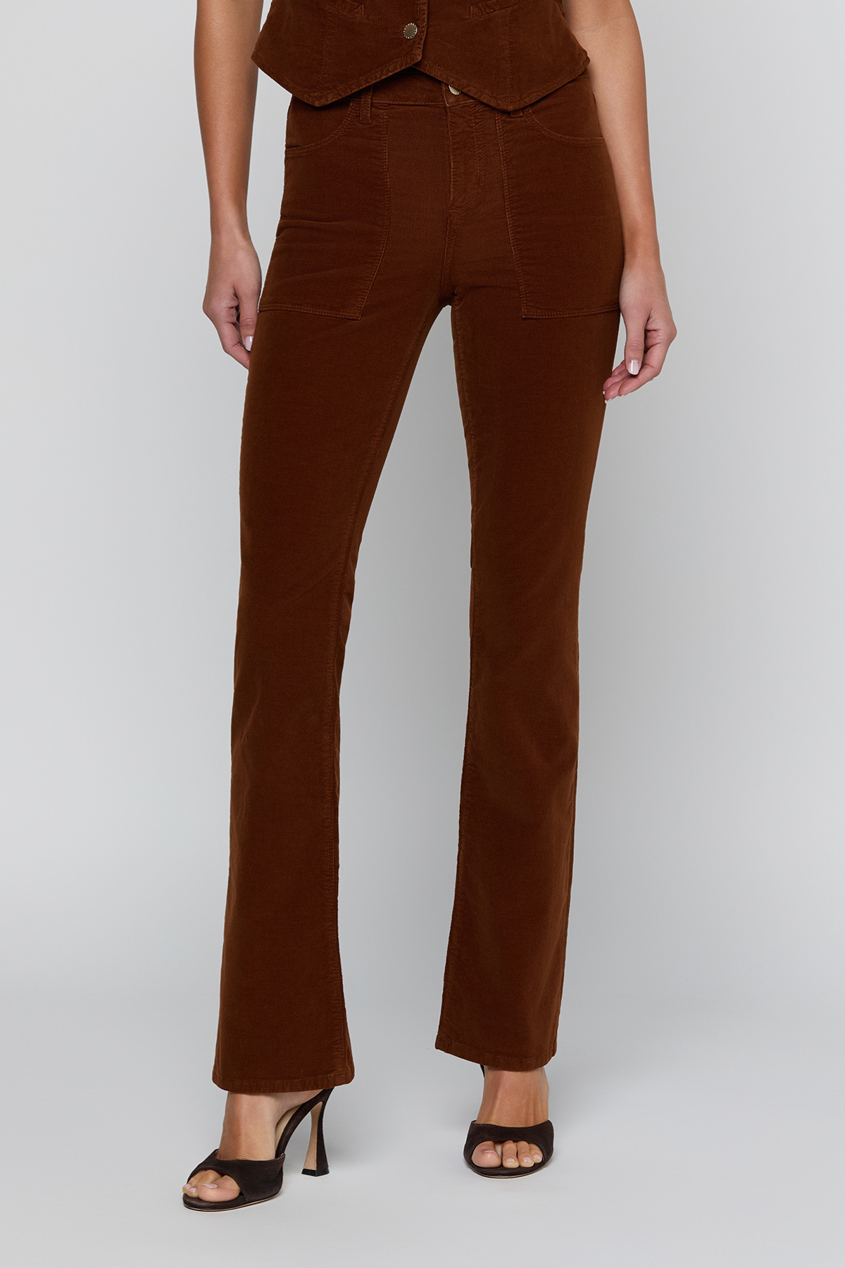 A model wearing brown corduroy flare pants with a matching brown vest and black heeled sandals, shown from the waist down against a light gray studio background.