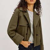 A woman wearing an olive green cropped double-breasted jacket over a light gray t-shirt and dark blue jeans, photographed against a neutral light gray studio background.