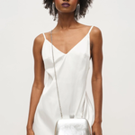 Woman wearing a white dress with feather details and holding a silver handbag against a plain background