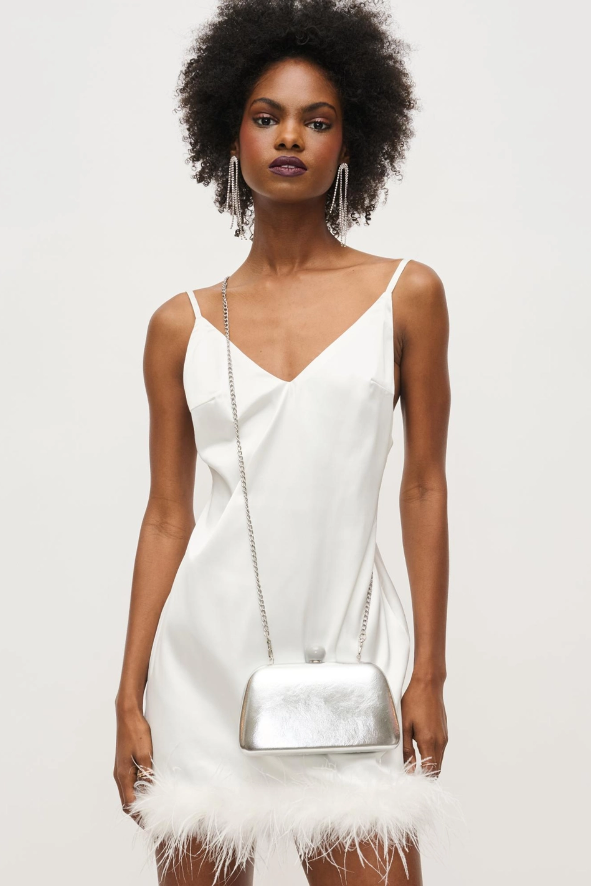 Woman wearing a white dress with feather details and holding a silver handbag against a plain background